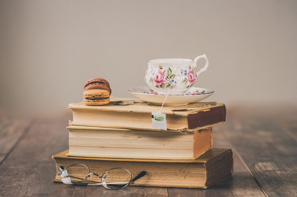 A pile of books, with a spectables, a cup of tea and biscuits beside it