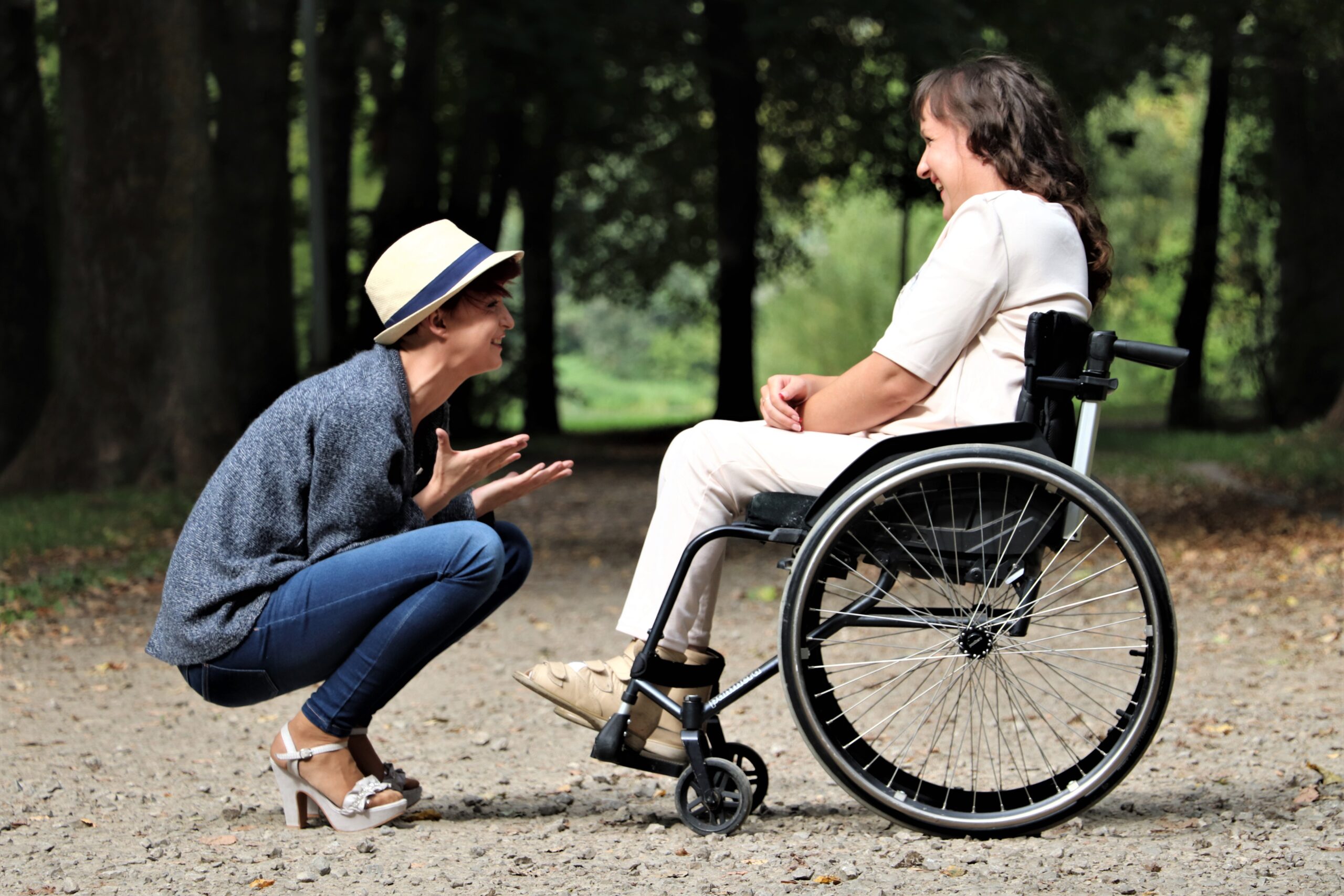 A woman on a wheelchair and another woman sitting chatting opposite her on the ground, to be on eye-level with the woman in the wheelchair.